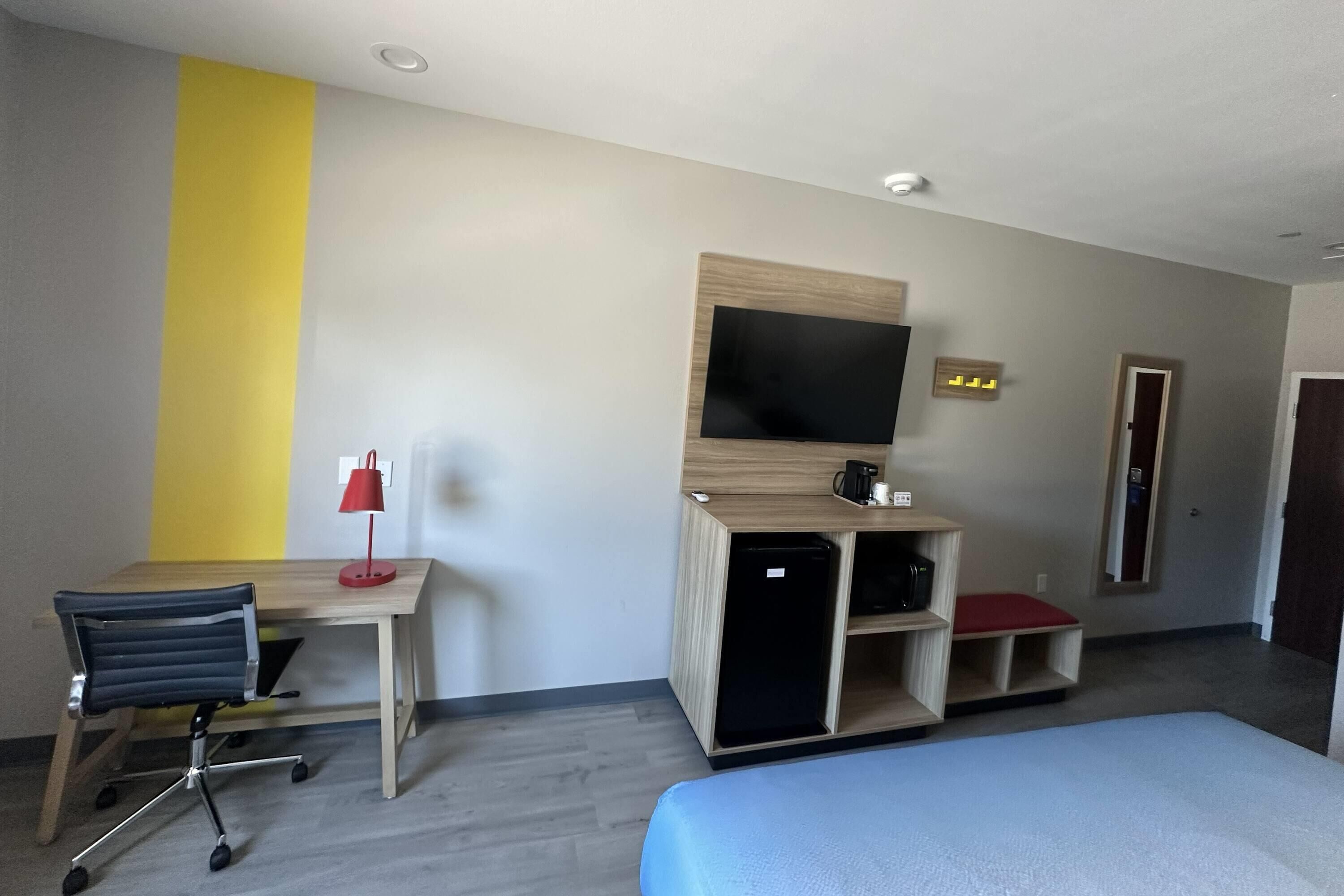 A hotel room with a bed, a desk with a chair, a television, and a small seating area with a red cushion.