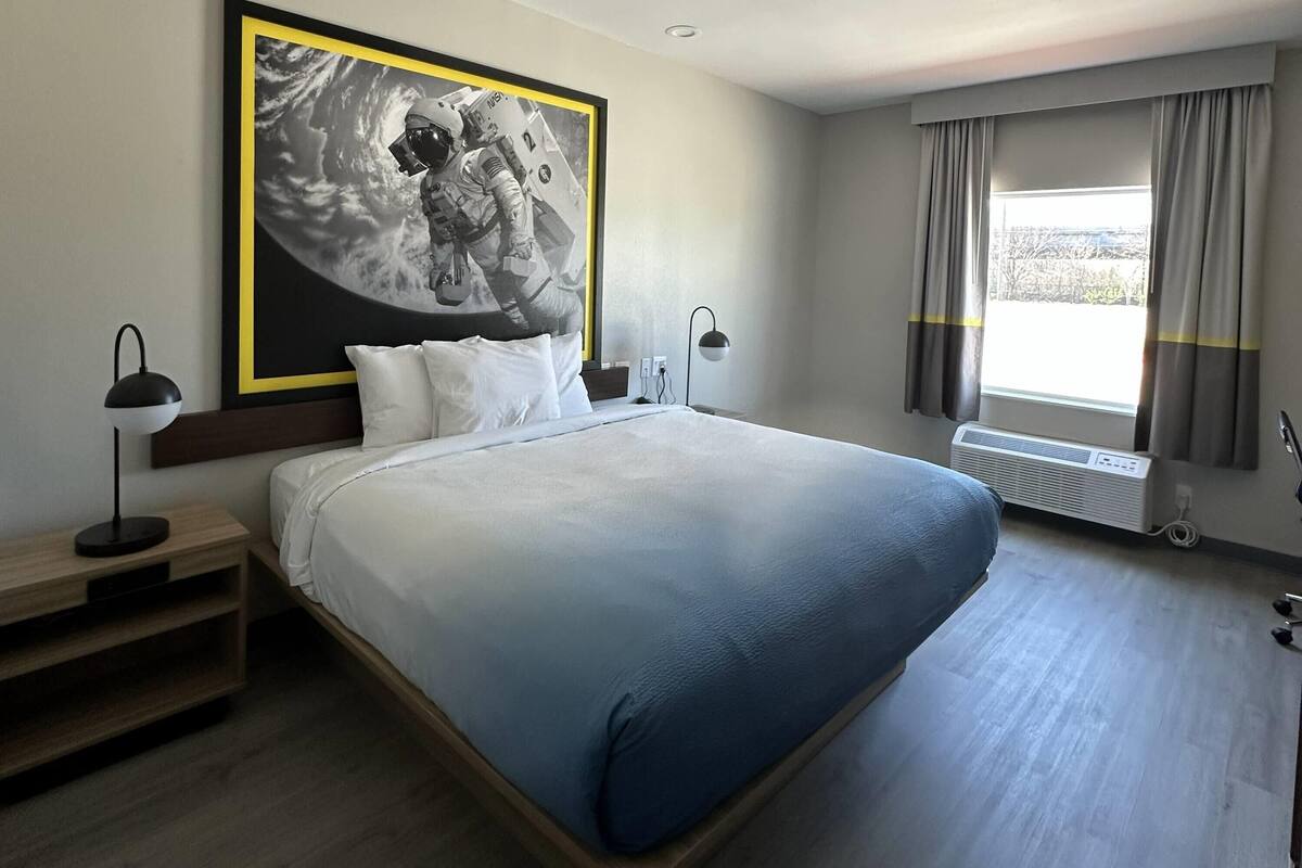 Una habitación de hotel con una cama grande, una mesita de noche con lámpara, un aire acondicionado instalado en la pared y una ventana con cortinas.