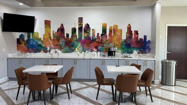 Una cocina moderna con un amplio mural de un paisaje urbano colorido, un televisor de pantalla plana y un comedor con sillas marrones y una mesa blanca.