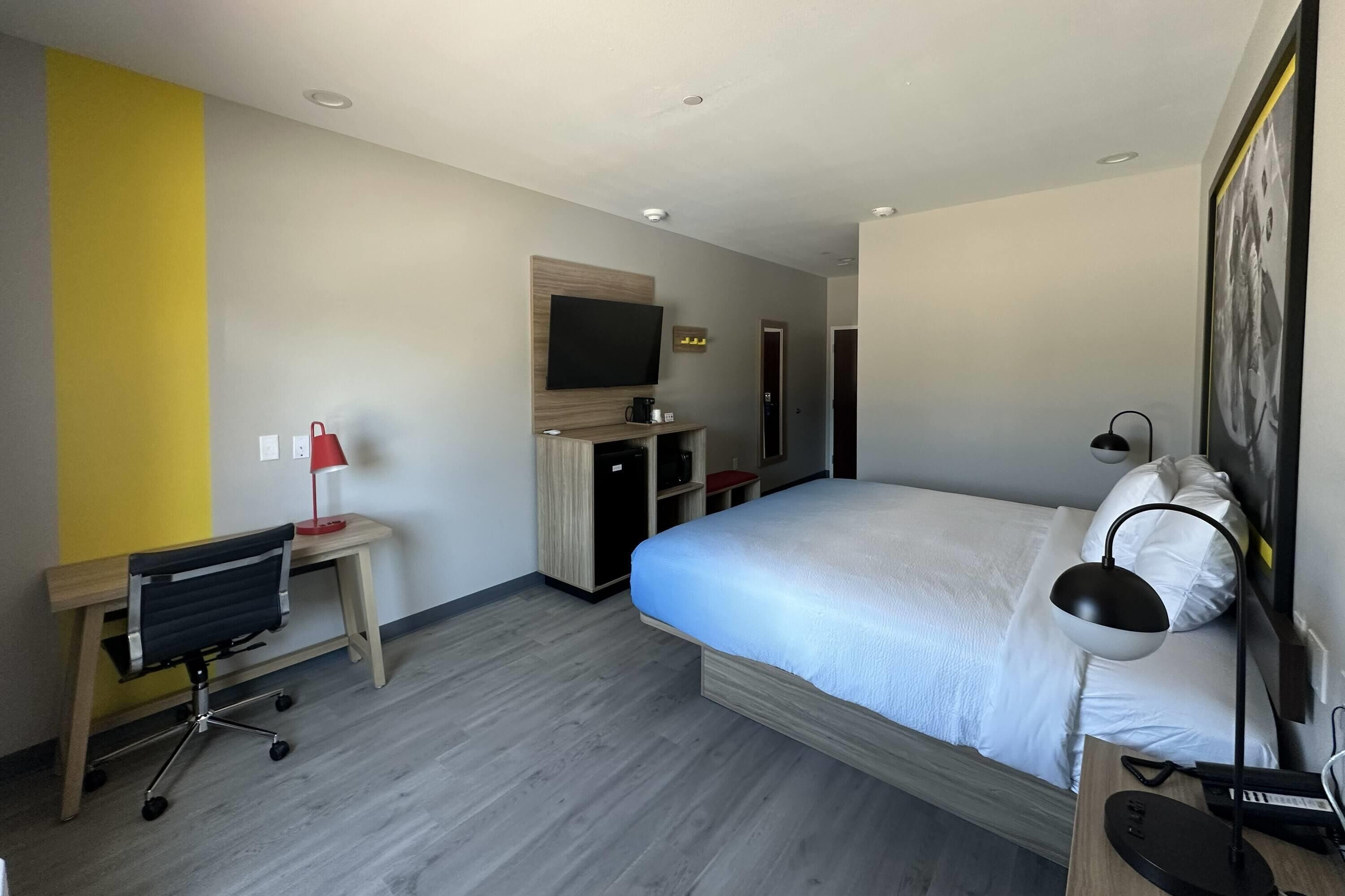 A modern hotel room with a bed, desk, chair, TV, and a small kitchenette.