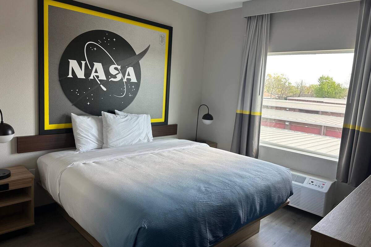 Habitación de hotel con un cuadro temático de la NASA, una cama con ropa de cama blanca, una mesita de noche con una lámpara y vistas a un edificio a través de una ventana.