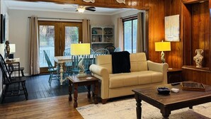 A living room with a wooden floor, a beige sofa, a wooden coffee table, and a dining area with a table and chairs. - The Fish Cottage - south beach, lots of amenities (Tybee Island)