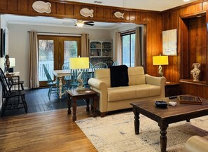 A living room with a wooden floor, a beige sofa, a wooden coffee table, and a dining area with a table and chairs. - The Fish Cottage - south beach, lots of amenities (Tybee Island)