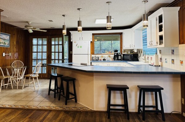 A kitchen with a large island, bar stools, a refrigerator, and a window with a view of trees. - The Fish Cottage - south beach, lots of amenities (Tybee Island)