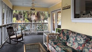 A covered porch with a wooden rocking chair, a floral sofa, a small table, and a ceiling fan. - The Fish Cottage - south beach, lots of amenities (Tybee Island)