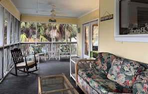 A covered porch with a wooden rocking chair, a floral sofa, a small table, and a ceiling fan. - The Fish Cottage - south beach, lots of amenities (Tybee Island)