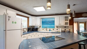 Una cocina con una isla grande, armarios blancos, un cuadro del océano e iluminación colgante.