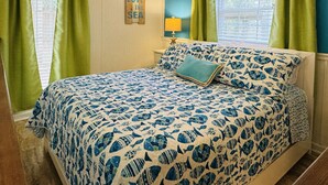Ein Schlafzimmer mit einem Bett, grünen Vorhängen, einem Deckenventilator, einer Wand mit dem Schild 'Love the Sea' und einem Fenster mit Jalousien.