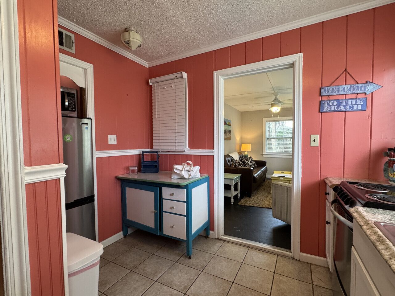 A compact kitchen with a refrigerator, microwave, and stove. A small table with drawers is against the wall, and a door leads to a living area.