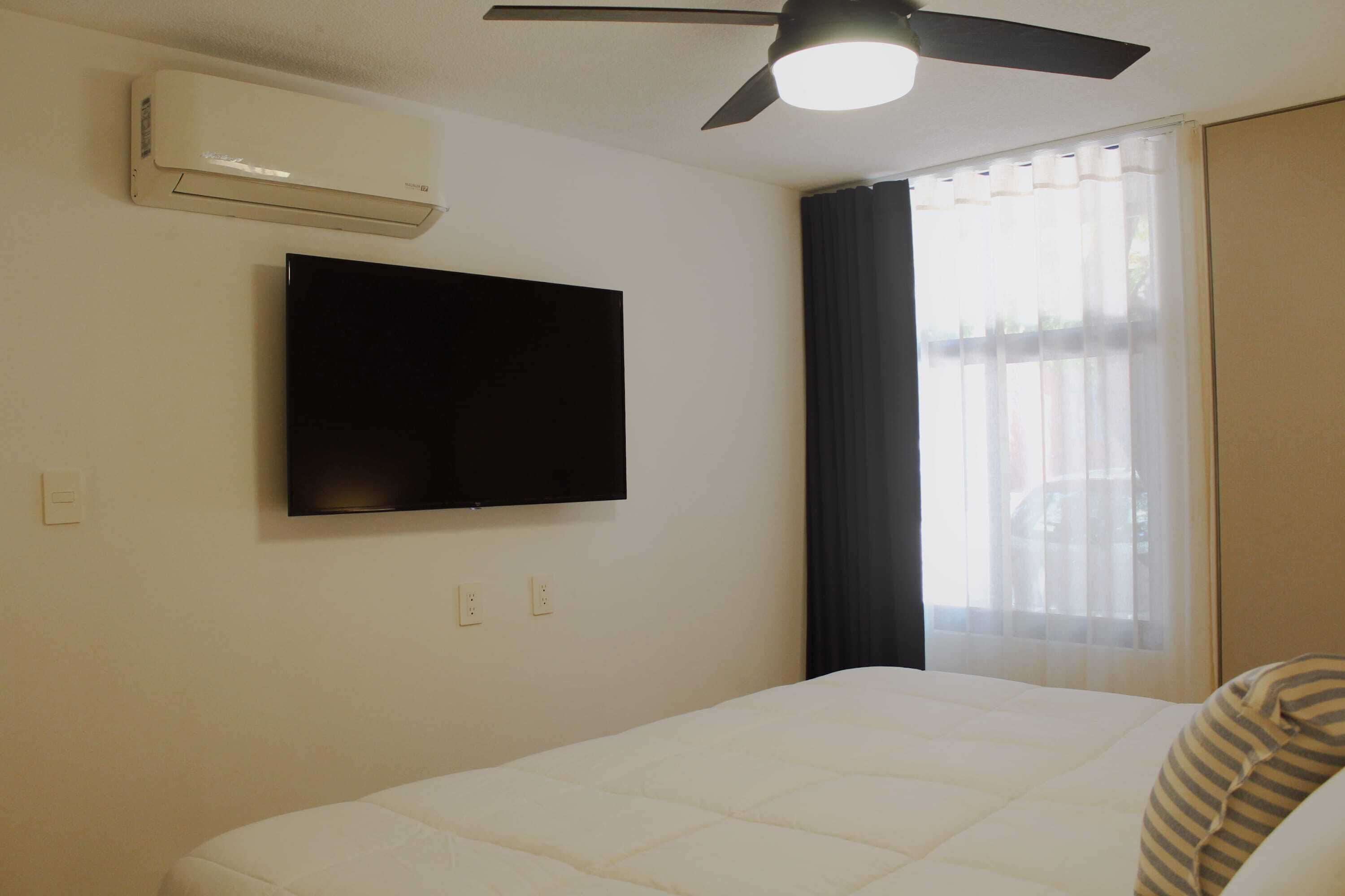 A hotel room with a bed, a flat-screen TV mounted on the wall, an air conditioner, a ceiling fan, and a window with sheer curtains.