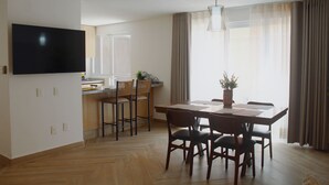 Ein modernes Wohnzimmer mit Flachbildfernseher an der Wand, einem Essbereich mit Holztisch und Stühlen sowie einer Küchenzeile inklusive Arbeitsfläche und Sitzgelegenheiten.