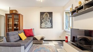 A modern living room with a grey sofa, a patterned rug, a flat-screen TV, and a wall-mounted shelf with decorative items. - MV - Three-room apartment, covered garage 15 minutes from downtown (Trieste)