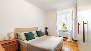 A bedroom with a wooden bed, a nightstand, a dresser, and a window with curtains. - MV - Three-room apartment, covered garage 15 minutes from downtown (Trieste)