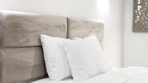 Uma cama com cabeceira, roupa de cama branca e luminária fixada na parede.