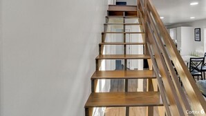 Uma escadaria de madeira com painéis de vidro que leva a um quarto com teto branco e uma área de jantar ao fundo.