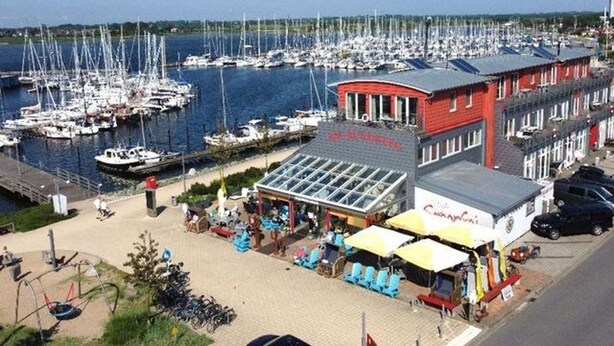 A marina with boats docked, a red building with a sign, outdoor seating, and a clear sky. - Apartment Fehmarn-burgtiefe (Fehmarn)