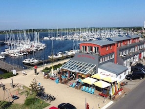A marina with boats docked, a red building with a sign, outdoor seating, and a clear sky. - Apartment Fehmarn-burgtiefe (Fehmarn)