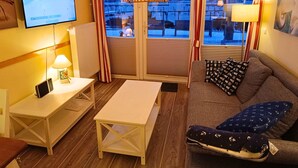A cozy room with a wooden floor, a sofa with patterned cushions, a small table, a TV on the wall, and a window with curtains. - Apartment Fehmarn-burgtiefe (Fehmarn)