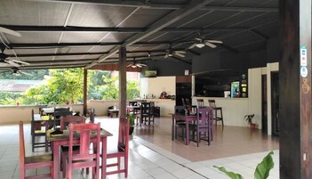 An open-plan restaurant with wooden tables and chairs, a bar area, and a view of greenery outside.