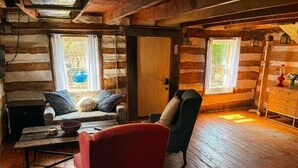 Interior de una cabaña de troncos con un sofá, sillón, mesa de madera y una puerta que da al exterior.