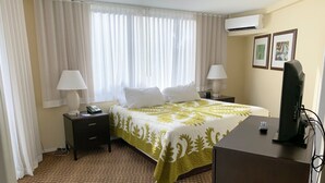 Une chambre d’hôtel comprenant un lit, deux tables de chevet avec des lampes, un bureau avec un ordinateur, un climatiseur et deux tableaux encadrés accrochés au mur.
