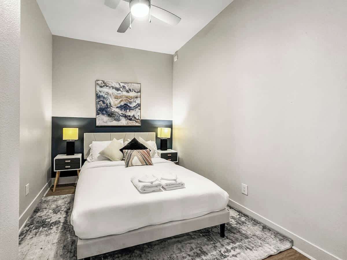 A modern hotel room with a large bed, two bedside tables with lamps, a ceiling fan, and a painting on the wall.