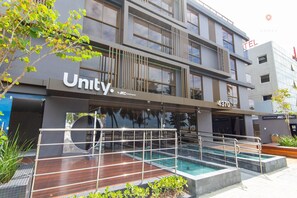 Een modern gebouw met een Unity hotelbordes, een zwembadgedeelte met een houten terras en een balkon met metalen hekwerk.