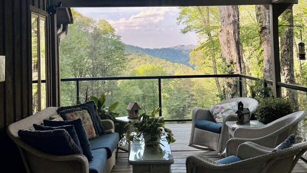 A covered outdoor seating area with wicker furniture, a glass coffee table, and a view of a forested mountain range.