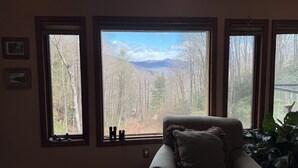 A room with a large window offering a view of a forested area and mountains. There is a chair with a cushion and a plant to the right.