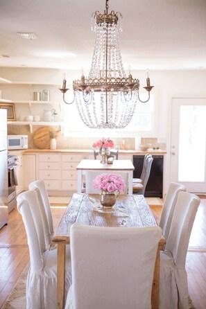 A dining area with a chandelier, white chairs, a wooden table, and a vase of pink flowers. - Perfect Round Top Girls Getaway | 2 BR w/ Fire Pit (Burton)