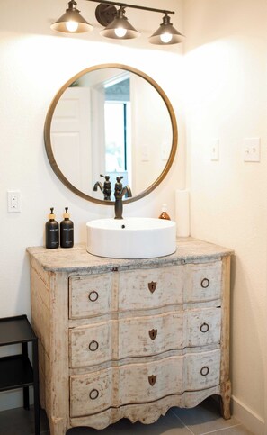 A bathroom with a white sink, a round mirror, and three pendant lights. There are two bottles of hand soap and a bottle of lotion on the sink. - Charming 2BR + Firepit | Minutes to Town + Shops (Burton)