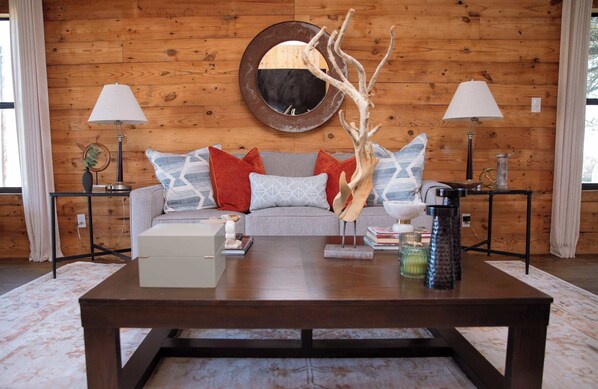 A cozy living room with a wooden wall, a round mirror, a sofa with patterned cushions, a coffee table with decorative items, and two lamps on side tables. - Charming 2BR + Firepit | Minutes to Town + Shops (Burton)