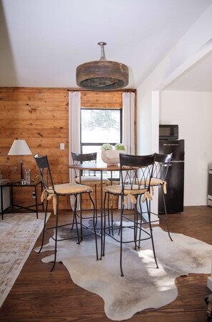 A dining area with a wooden wall, a chandelier, a table with chairs, a refrigerator, and a window with curtains. - Charming 2BR + Firepit | Minutes to Town + Shops (Burton)