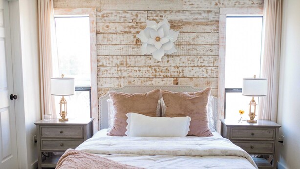 Een slaapkamer met een rustieke houten wand, een groot bed met veel kussens, twee nachtkastjes met lampen en een raam met gordijnen.
