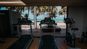 Un gimnasio con cintas de correr y una máquina elíptica, con vistas a una playa con palmeras.