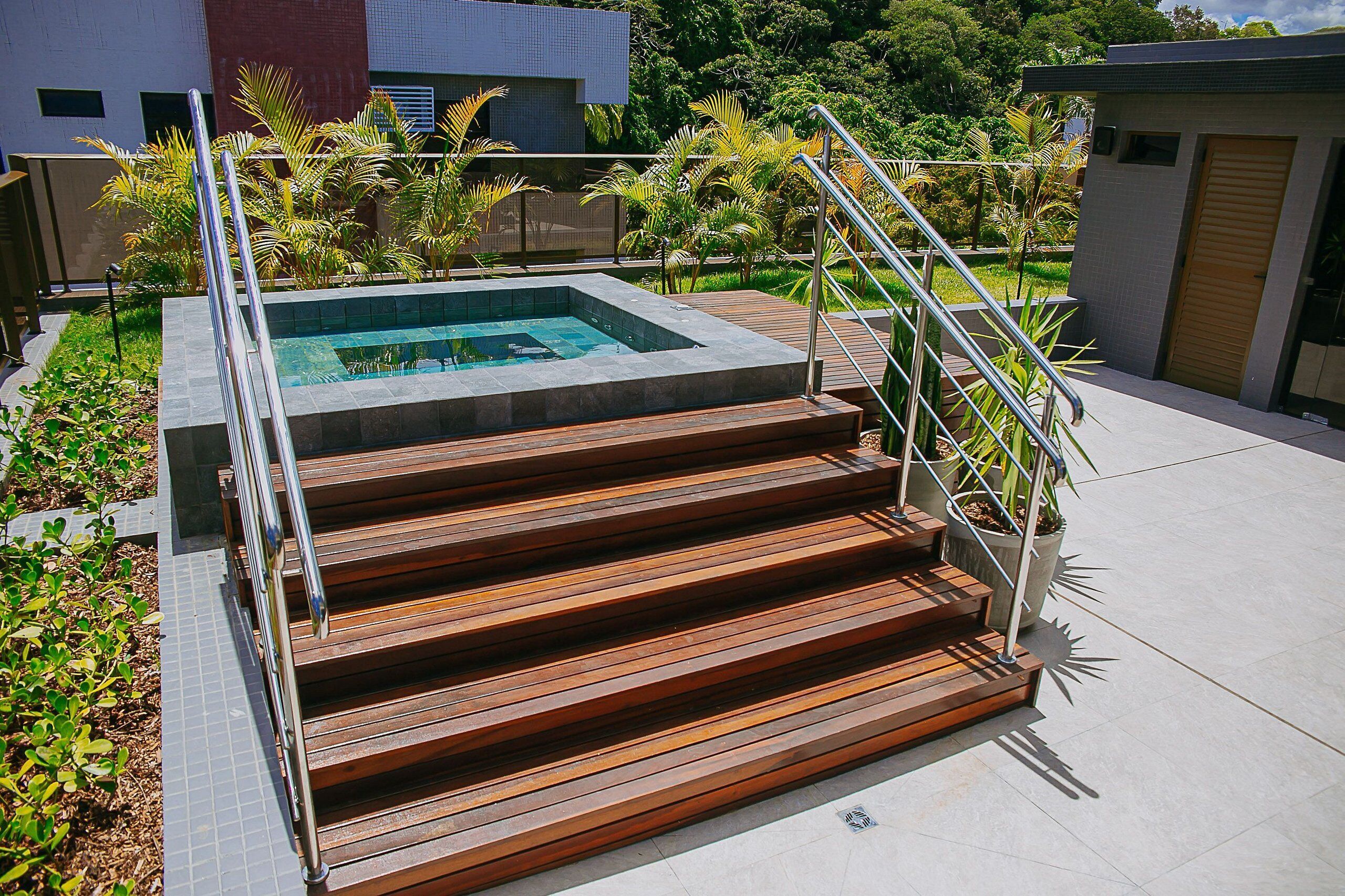 Uma área moderna de piscina ao ar livre com escadas de madeira que levam a piscina, cercada por vegetação exuberante e um guarda-corpo de metal.