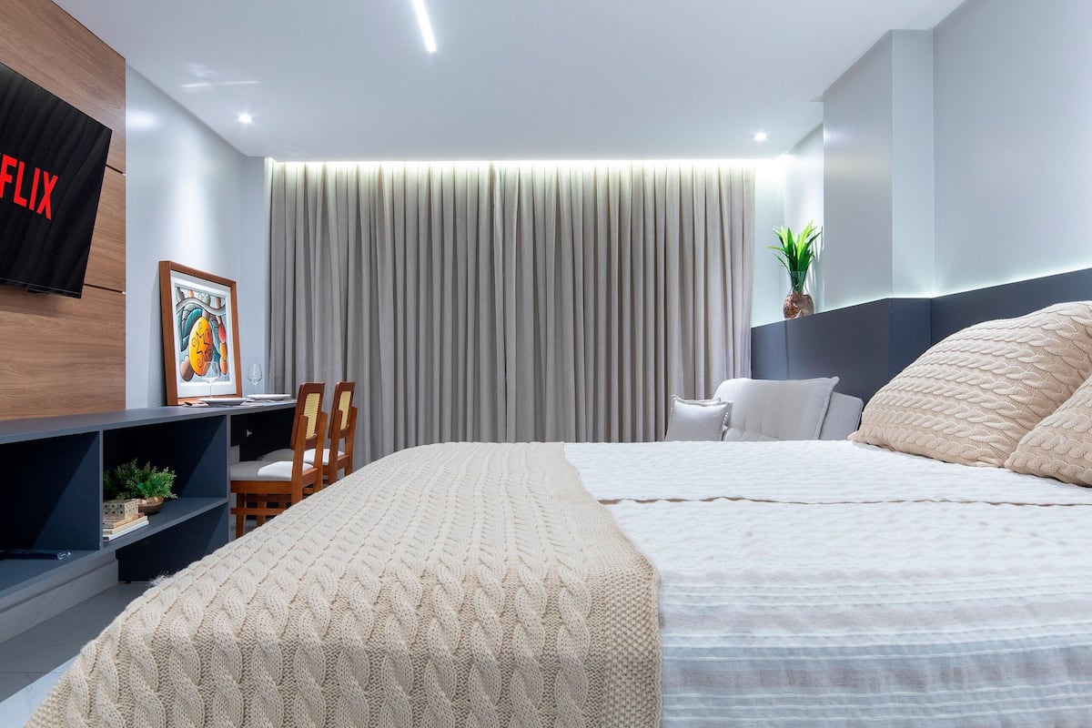 A modern bedroom with a bed, a TV mounted on the wall, a bookshelf, and a large window with curtains.