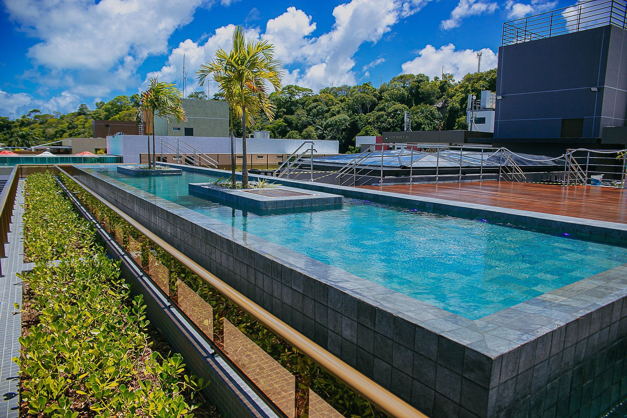 A swimming pool with a tiled edge, a wooden deck, and a view of buildings and trees in the background.
