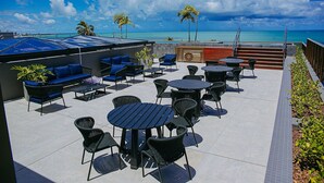 Een terras buiten met zitplaatsen en tafels, met uitzicht op een strand met palmbomen en de oceaan.