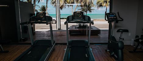 A gym with treadmills and an exercise bike, overlooking a beach with palm trees.