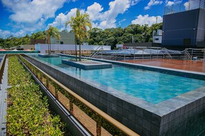 Ein Schwimmbecken mit einer gefliesten Kante, einer Holzterrasse und Blick auf Gebäude und Bäume im Hintergrund.
