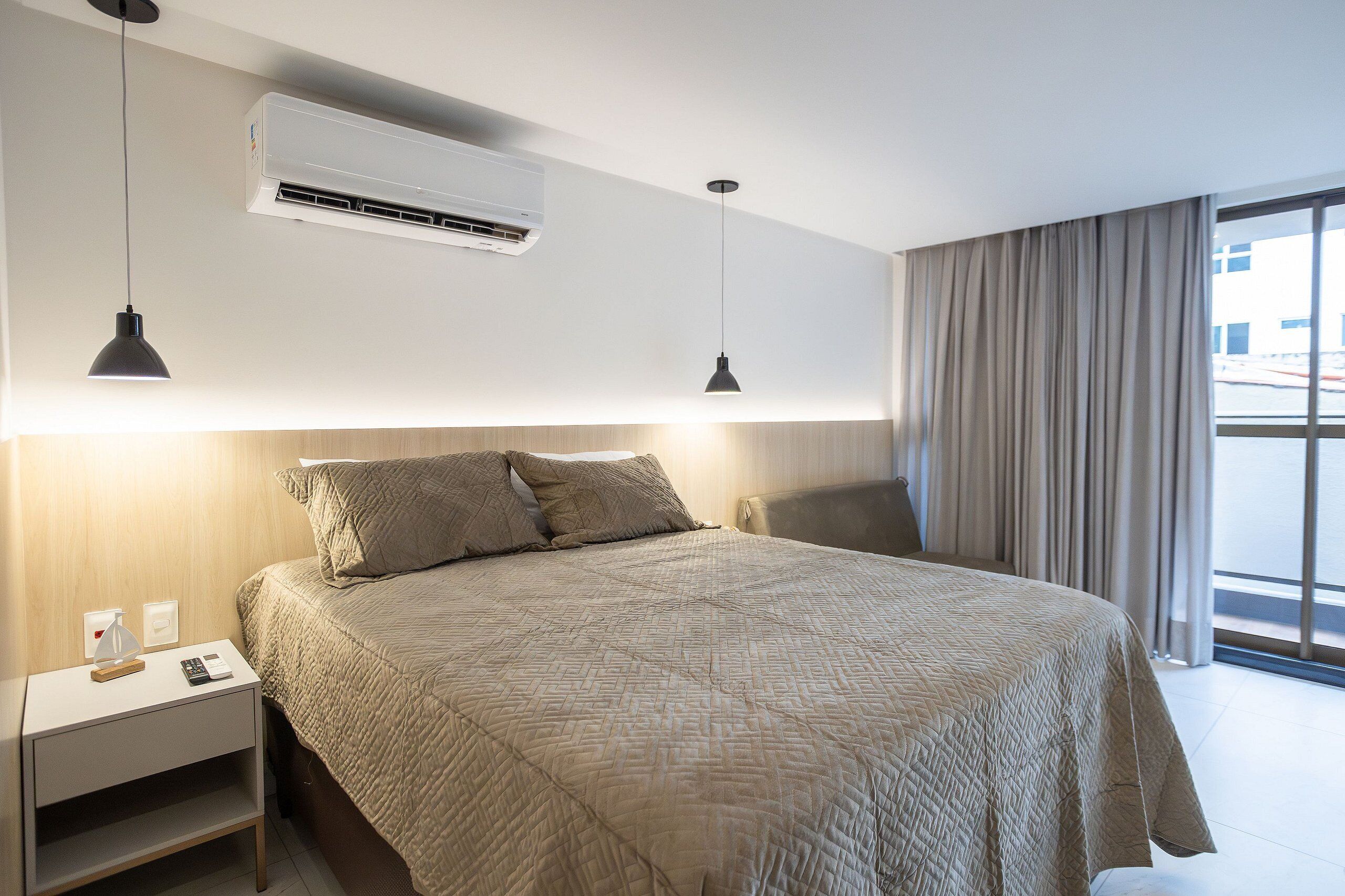 Una camera d'albergo moderna con un letto grande, due lampade a sospensione, un condizionatore d'aria, un comodino con una lampada e vista all'esterno tramite un'ampia finestra.