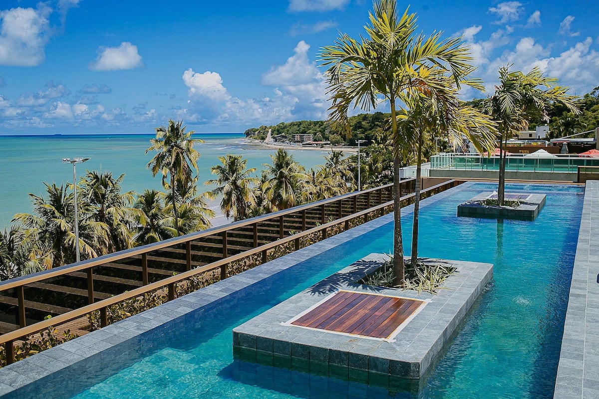 A swimming pool with a wooden deck, surrounded by palm trees and overlooking a beach.