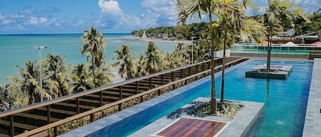 Una piscina con terraza de madera, rodeada de palmeras y vista a una playa.
