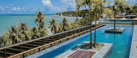 Zona de piscina con terraza de madera, palmeras y vista al océano.