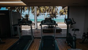 Ein Fitnessraum mit Laufbändern und Fahrrädern mit Blick auf einen Strand und Palmen.