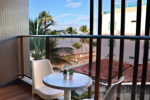 Ein Balkon mit einem weißen Stuhl, einem kleinen Tisch und zwei topfgepflanzten Pflanzen, mit Blick auf Palmen und Gebäude.