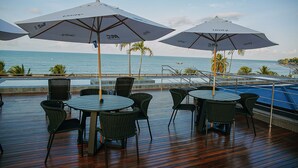 Een terras met tafels en stoelen, parasols en uitzicht op de oceaan.