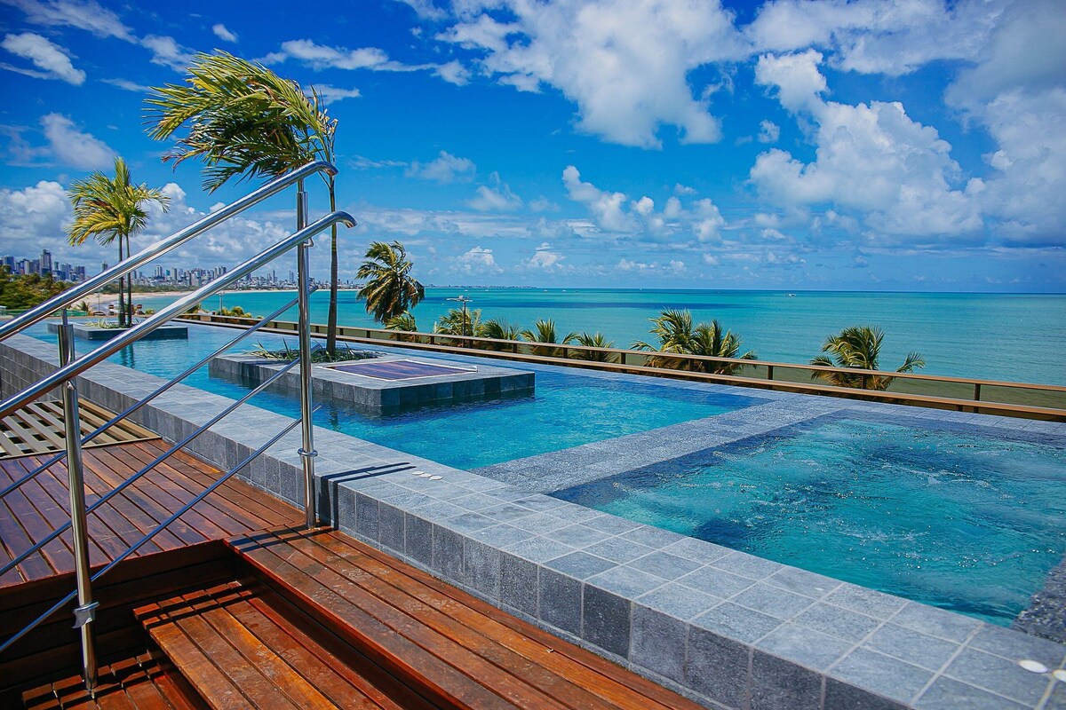 Ein Swimmingpool mit Holzterrasse und Metallgeländern, mit Blick auf das klare, blaue Meer und eine ferne Stadtlandschaft.