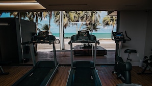 Un gimnasio con cintas de correr y una máquina elíptica, con vistas a una playa con palmeras.